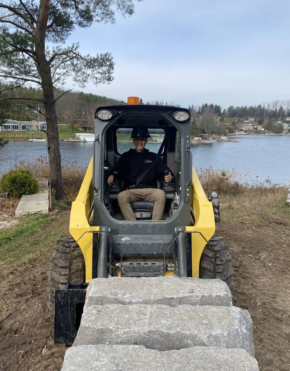 Skid steer loader hauling a large armour stone block at a lakeside job site