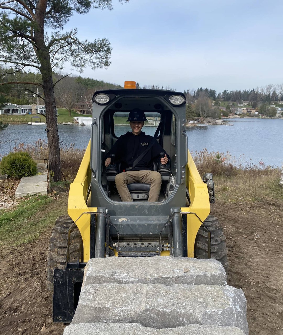 Skid steer loader hauling a large armour stone block at a lakeside job site